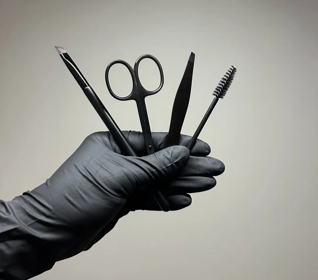 Wenkbrauwgereedschap in een gehandschoende hand: pincet, schaartje, borsteltje en potlood